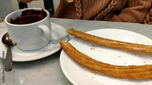 Female hand grabbing some churros from a plate and dipping it in a mug of thick dark hot chocolate.  The chocolate is thick and light shines on the surface.  Is tradition to have them on New Years Day photo