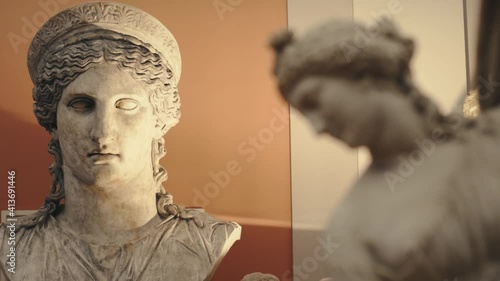 Ancient antique statue of a young woman in a museum with moody warm lights and a head of a bigger woman statue in the background. Smooth dolly sliding movement with parallax effect in 4K.