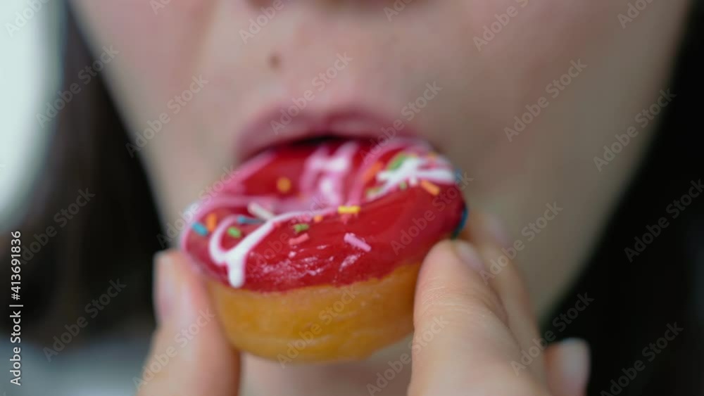 A woman pleasantly smiling bites a soft donut covered with red fruit glaze and confectionery sprinkles. Rich food. A woman pleasantly smiling bites a soft donut covered with red fruit glaze and confectionery sprinkles. Rich food.