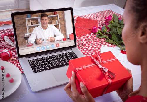 Diverse couple making valentine's date video call using laptop both holding gifts
