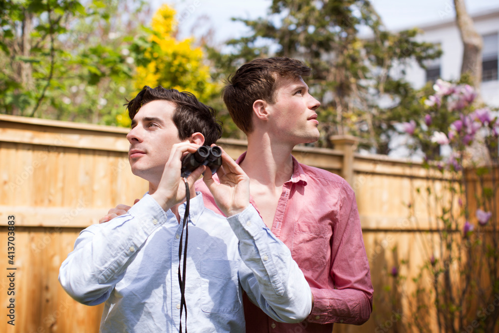 Gay men with binocular looking away while standing in backyard Gay men with binocular looking away while standing in backyard
