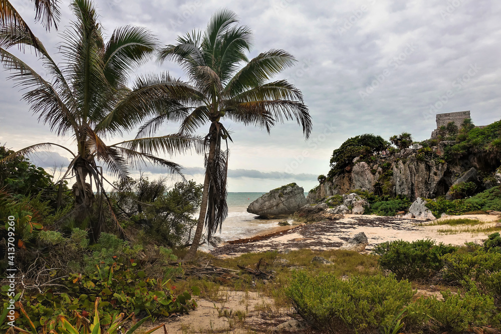 Beach in the Mayan city of Tulum. On the shore there are tropical vegetation, palms. There are stones and algae on the sand. The ruins of an ancient building are visible on the rocky shore. Mexico Beach in the Mayan city of Tulum. On the shore there are tropical vegetation, palms. There are stones and algae on the sand. The ruins of an ancient building are visible on the rocky shore. Mexico