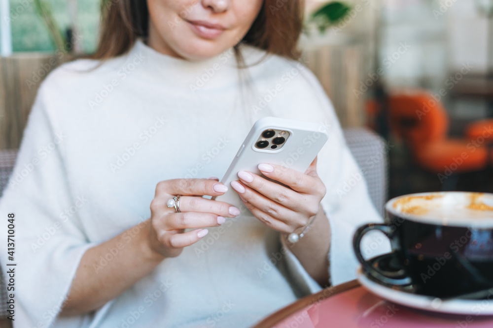 Fototapeta premium Attractive young brunette smiling woman in white casual dress with cup of coffee using mobile phone in cafe