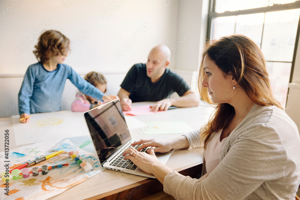 Woman using laptop computer while father assisting children in drawing at table Woman using laptop computer while father assisting children in drawing at table