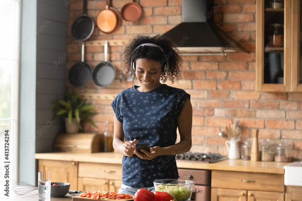 Smiling African American woman wearing headphones using phone, listening to music, enjoying leisure time, standing in kitchen, searching vegetarian snacks recipe or chatting, cooking salad at home - obrazy, fototapety, plakaty 