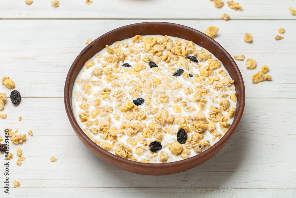 Healthy breakfast. Muesli with dried grapes is poured with milk in a clay bowl on a white wooden table. Muesli is scattered on the table. Healthy breakfast. Muesli with dried grapes is poured with milk in a clay bowl on a white wooden table. Muesli is scattered on the table.