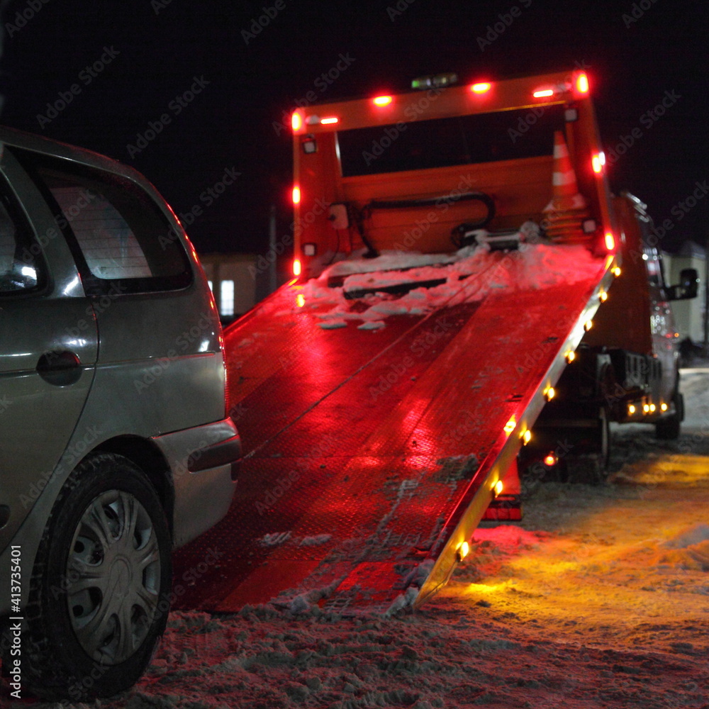 Tow truck loads a car on an inclined platform with a winch on a winter night on the city road, technical assistance service to the driver