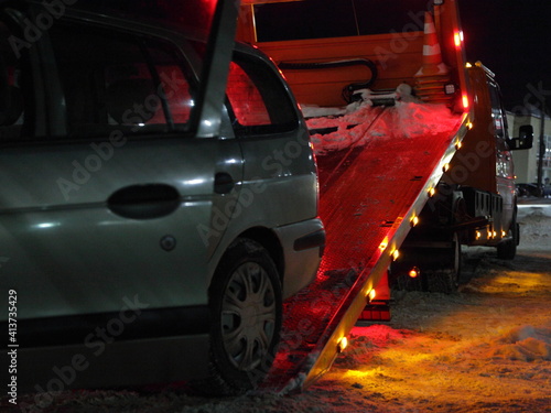 Tow truck loads a car with opened driver door onto a platform on a winter night on the city road, technical assistance to the driver