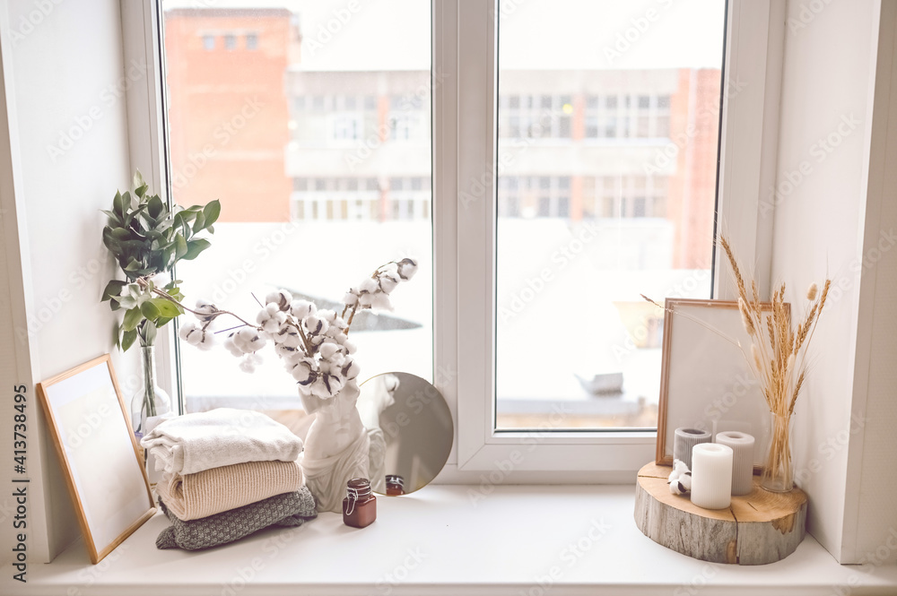 Cozy still life feminine scene pastel colors. Female styled window sill minimalistic composition. Empty mock up poster frame, elegant accessories mirror, folded sweaters, candles, dry cotton flowers.