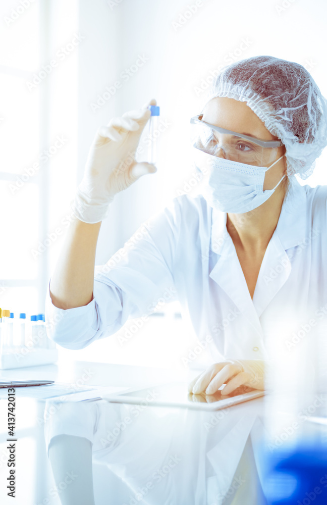 Professional female scientist in protective eyeglasses researching tube with reagents in sunny laboratory toned in blue. Medicine and science researching Professional female scientist in protective eyeglasses researching tube with reagents in sunny laboratory toned in blue. Medicine and science researching