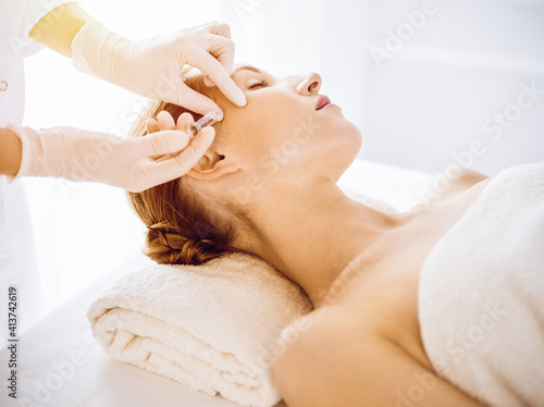 Beautiful woman receiving beauty injections with closed eyes in sunny medical center. Beautician doctor hands doing beauty procedure to female face with syringe. Cosmetic medicine and surgery concept