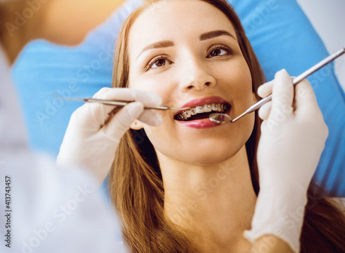 Smiling young woman with orthodontic brackets examined by dentist in sunny dental clinic. Healthy teeth and medicine concept