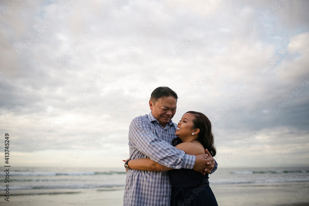 Happy woman embracing father while standing at beach against cloudy sky Happy woman embracing father while standing at beach against cloudy sky