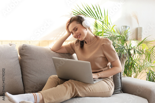 Smiling young woman with headphones and laptop on the sofa.
