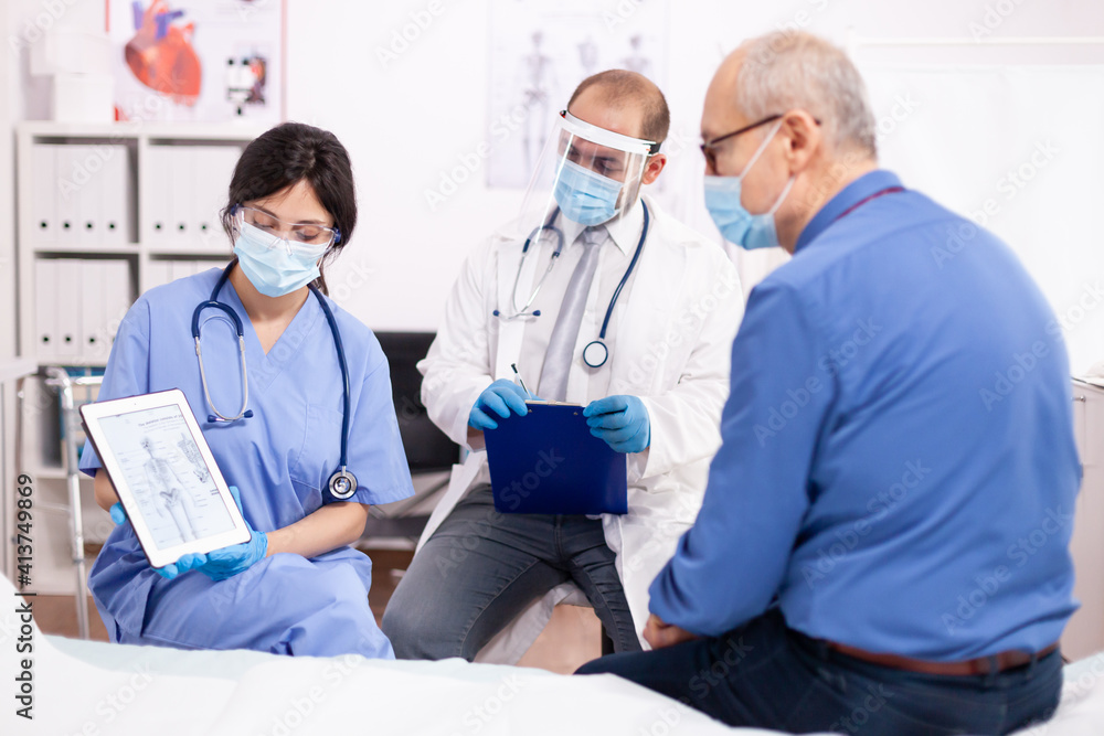 Doctor discussing about bone disease with senior man pointing at tablet pc in cardiology wearing face mask in time of covid19. Medical staff in uniform discussing about bone injury with patient during Doctor discussing about bone disease with senior man pointing at tablet pc in cardiology wearing face mask in time of covid19. Medical staff in uniform discussing about bone injury with patient during