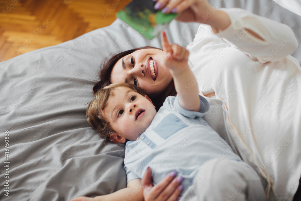 High angle view of happy mother lying with baby boy pointing at picture book on bed