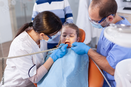 Little girl with mouth open in the course of cavity treatment sitting on dental chair. Mother with her kid in stomatology clinic for teeth examine using modern instruments.