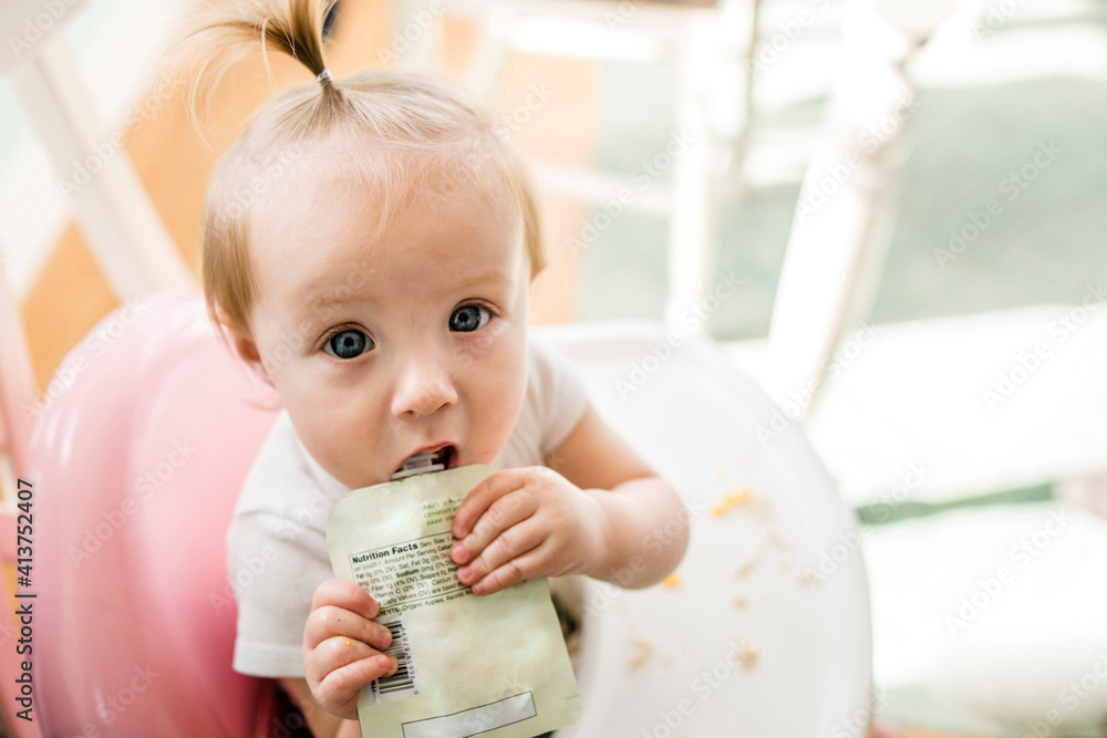 High angle portrait of cute baby girl eating apple sauce from pack while sitting on high chair at home High angle portrait of cute baby girl eating apple sauce from pack while sitting on high chair at home
