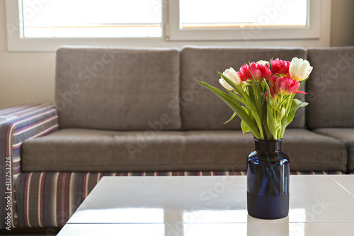 Close up shot of blue glass vase with peony tulips on white table on foreground and gray couch with colorful stripes under the window on background. Simple interior design. Copy space for text.