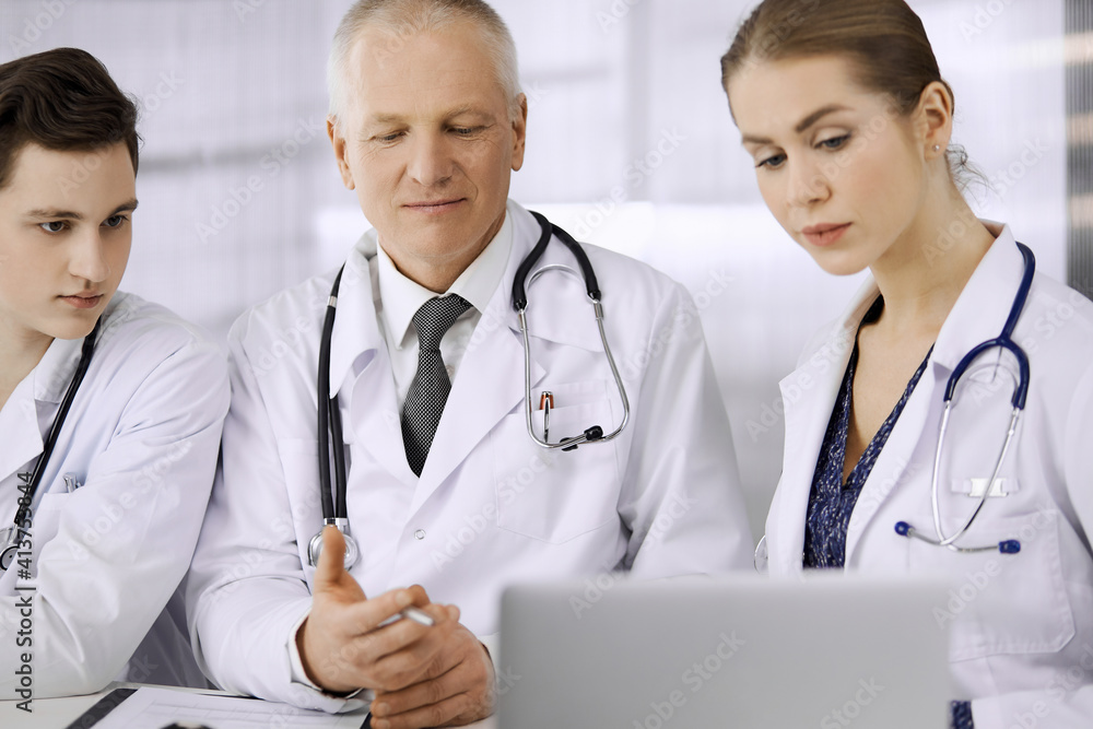Doctors colleagues and elderly therapist discussing current disease therapy while sitting at working place in clinic. Teamwork in hospital. Medicine and healthcare concept Doctors colleagues and elderly therapist discussing current disease therapy while sitting at working place in clinic. Teamwork in hospital. Medicine and healthcare concept