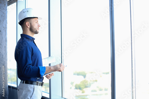 Confident young business man in shirt examining blueprint while standing against a window at office.