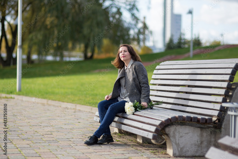 A woman in a coat sits on a bench in a sunny autumn park. Nearby lies a bouquet of white roses, she smiles and looks away. A woman in a coat sits on a bench in a sunny autumn park. Nearby lies a bouquet of white roses, she smiles and looks away.