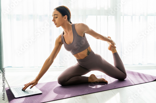 Enduring concentrated sporty woman is doing working out at home and doing plank in front of her laptop, wearing sport outfit