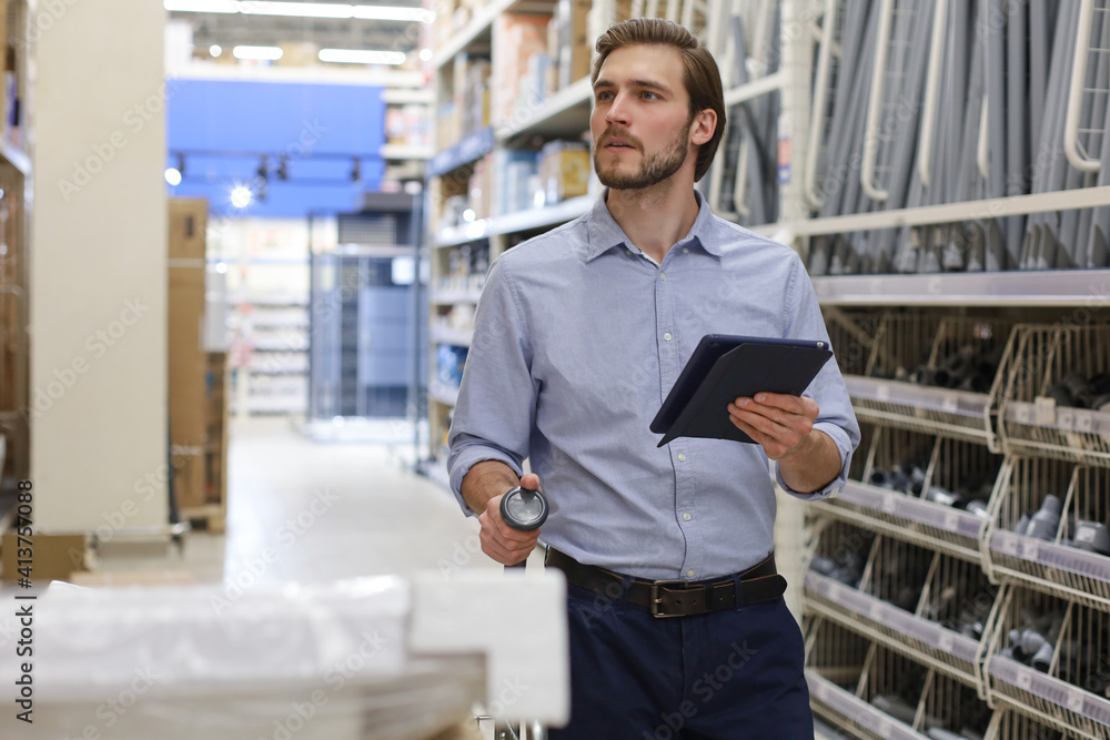 Young man shopping or working in a hardware warehouse standing checking supplies on his tablet. Young man shopping or working in a hardware warehouse standing checking supplies on his tablet.