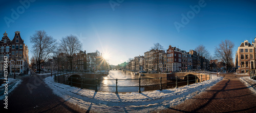 Wide panorama image of the Amsterdam canals at the Leidsegracht with snow and ice during golden hour on a beautiful sunny day with blue sky