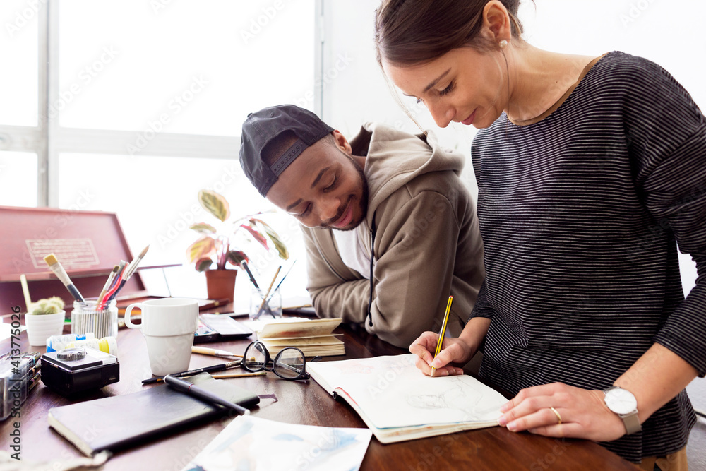 Smiling male illustrator looking at female colleague painting at desk in creative office Smiling male illustrator looking at female colleague painting at desk in creative office