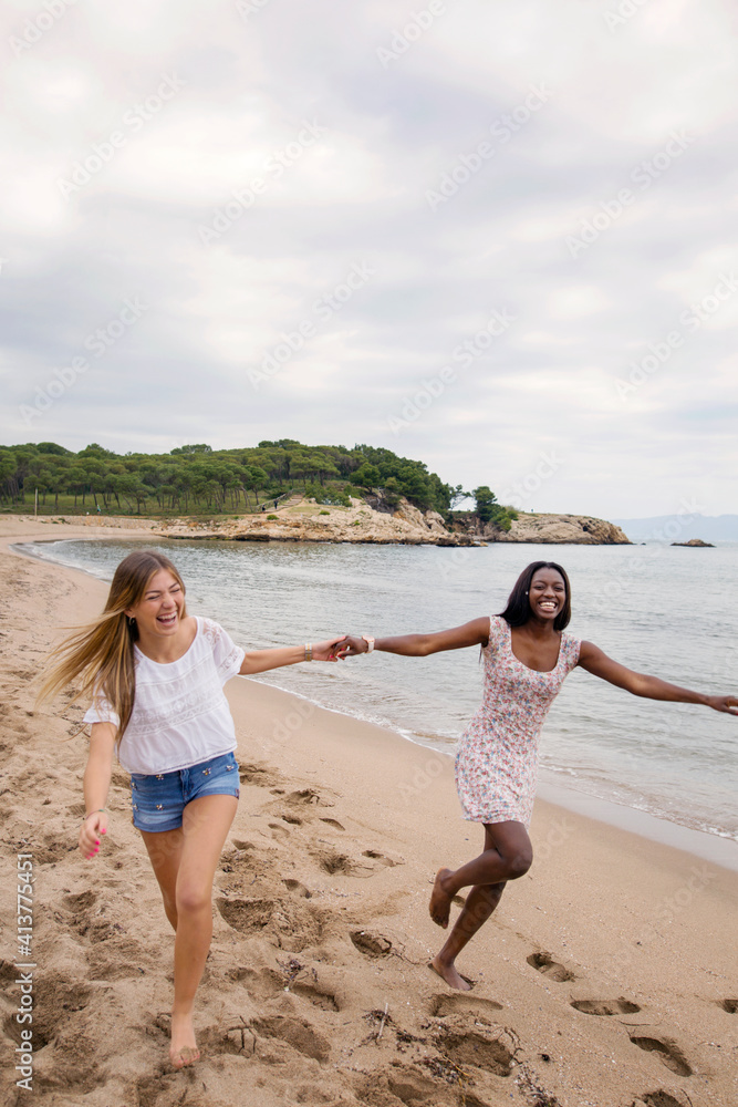 Cheerful female friends holding hands while running on beach against sky Cheerful female friends holding hands while running on beach against sky