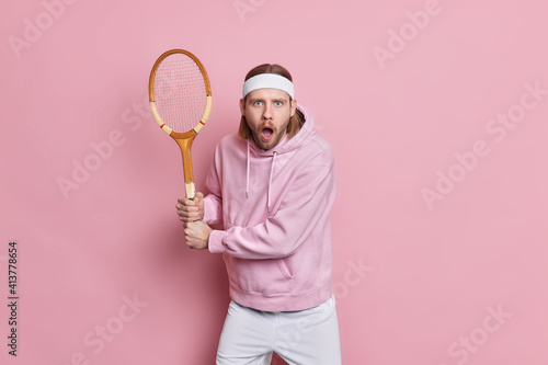 Shocked handsome male tennis player poses in ready position with racquet waits for serve dressed in sportswear being very attentive while playing poses indoor against pink studio background. © Wayhome Studio