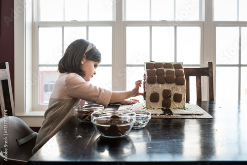 Side view of girl making gingerbread house on table against window at home photo