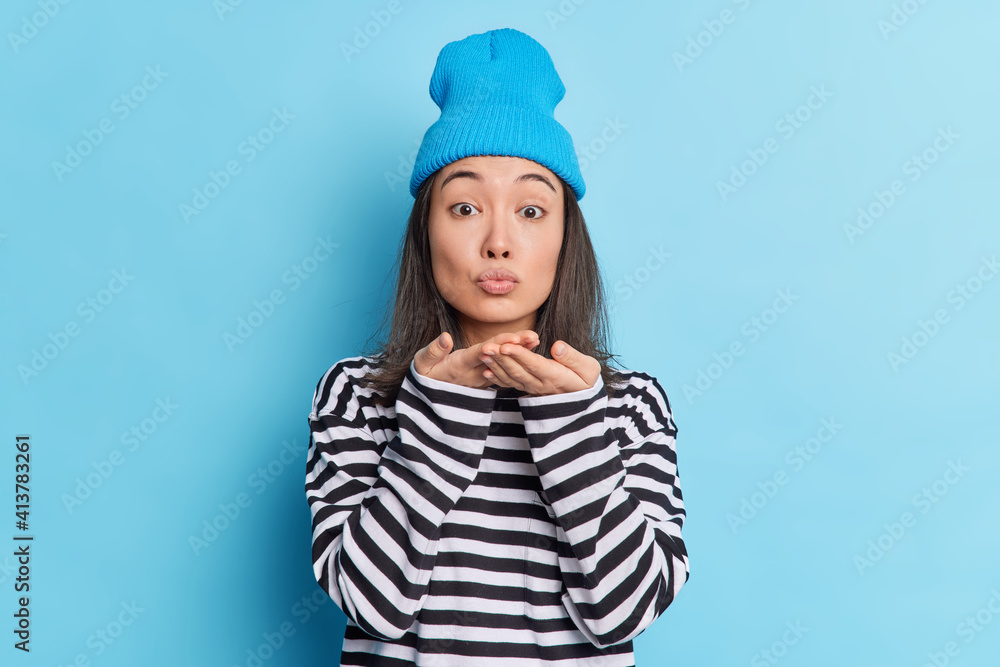 Studio shot of lovely Asian woman blows airkiss keeps lips folded palms near mouth expresses love to boyfriend dressed in stylish outfit isolated on blue background. Affectionate girlfriend gives mwah Studio shot of lovely Asian woman blows airkiss keeps lips folded palms near mouth expresses love to boyfriend dressed in stylish outfit isolated on blue background. Affectionate girlfriend gives mwah