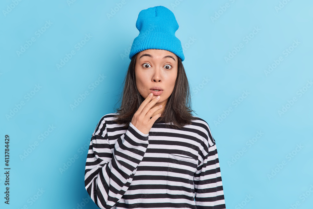 People emotions reaction concept. Stunned Asian girl stares at camera holds breath sees somethnign unexpected and shocking dressed in striped jumper stylish hat isolated on blue studio wall. People emotions reaction concept. Stunned Asian girl stares at camera holds breath sees somethnign unexpected and shocking dressed in striped jumper stylish hat isolated on blue studio wall.