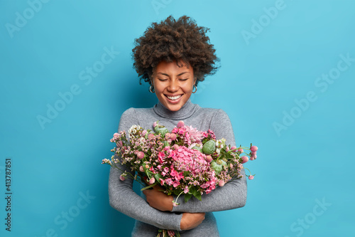 Lovely Afro American woman embraces bouquet gets spring flowers on Womans Day expresses positive emotions dressed casually isolated over blue background. Festive event and celebration concept