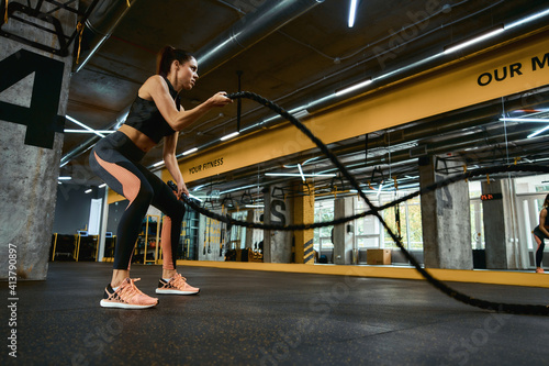 Cardio workout. Young strong athletic woman wearing sport clothes exercising with battle ropes at crossfit gym, full length