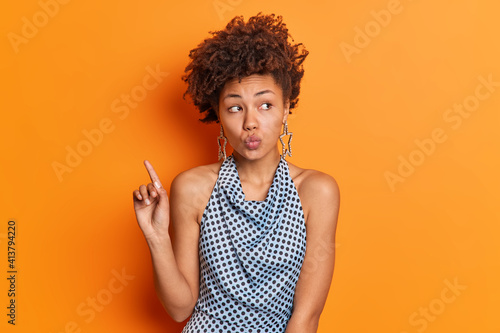 Glamour lovely African American female model keeps lips rounded indicates above gazes aside shows some product dressed in fashionable outfit poses against vivid orange background. Look upwards