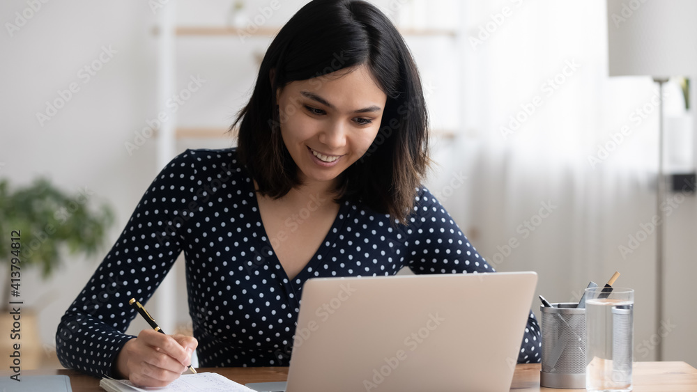 Asian female college university student watch tele lecture on pc make notes of training course. Concentrated young woman translator handwriting details translating video material to english language Asian female college university student watch tele lecture on pc make notes of training course. Concentrated young woman translator handwriting details translating video material to english language