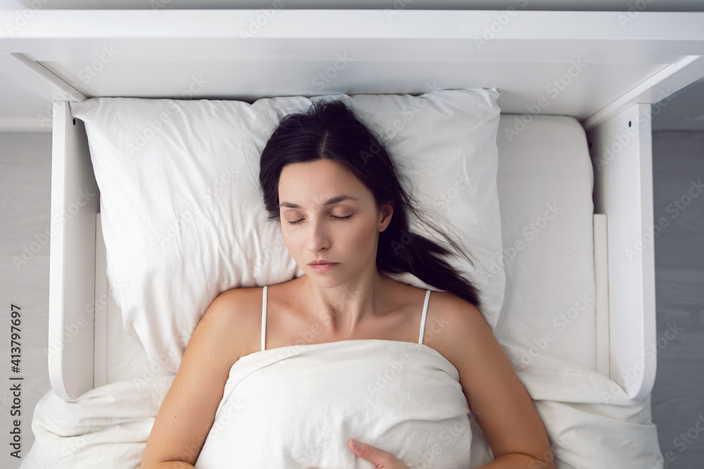 woman in a white t-shirt is lying on the bed and a pillow is under the blanket in the room