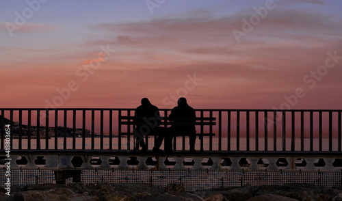 two silhouettes of men talking sitting on a bench watching the sunset on the beach 