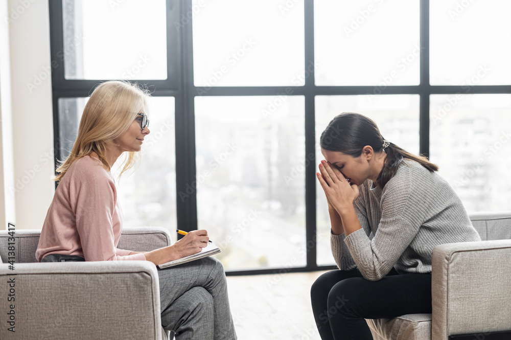 Girl at a psychologist's appointment,talking about hard period,holding her hands near the head,crying,blonde woman writing down notes,the psychologist with glasses is listening attentively,advising Girl at a psychologist's appointment,talking about hard period,holding her hands near the head,crying,blonde woman writing down notes,the psychologist with glasses is listening attentively,advising