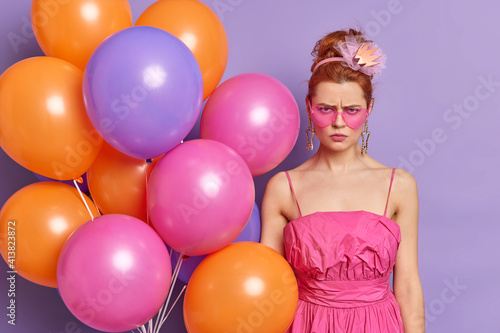 Offended displeased redhead woman looks angrily at camera wears pink shades and dress dissatisfied with celebration poses on party with multicolored balloons isolated over purple background. © wayhome.studio 