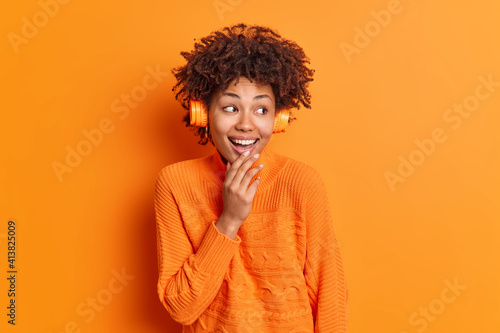 Positive dark skinned young woman with curly hair listens audio track wears wireless headphones looks gladfully aside dressed in casual jumper isolated over orange background enjoys nice sound