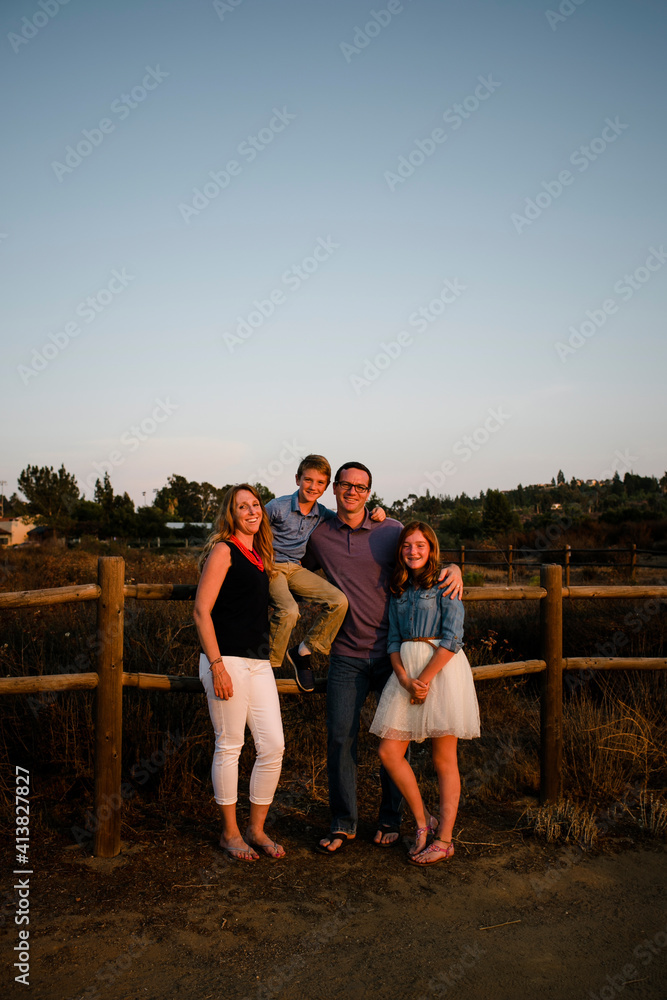 Full length portrait of happy family by fence on field against sky during sunset Full length portrait of happy family by fence on field against sky during sunset