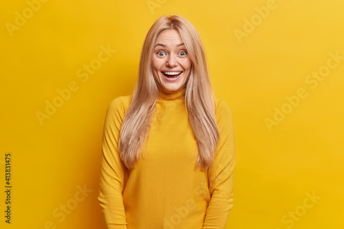 Positive blonde woman smiles broadly reacts on awesome news stares gladfully at camera wears casual turtleneck isolated over yellow background. Happy emotions feelings and gladfulness concept
