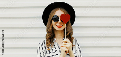 Portrait of happy smiling woman covering her eye with red heart shaped lollipop on a white background
