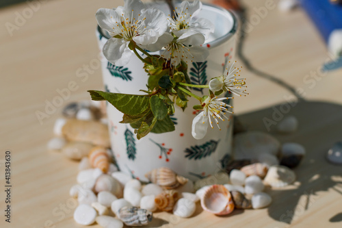 White candle with patterns on a beige table with sea stones, shells and a flowering cherry branch on a sunny day side view