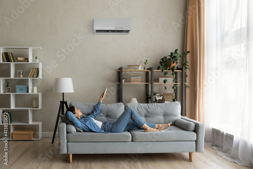 Glad millennial female buyer of air conditioner relax on sofa hold controller breath cool fresh air despite of hot summer day outside. Happy young lady regulate climate at home using modern ac device photo