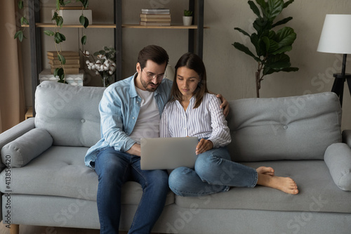 Serious young couple sit on sofa by laptop consider on making expensive purchase discuss wedding planning. Loving millennial spouses surf internet together search for new house apartment to buy rent
